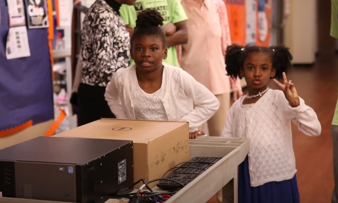Two girls at a cart loaded with a donated computer, one giving a peace sign