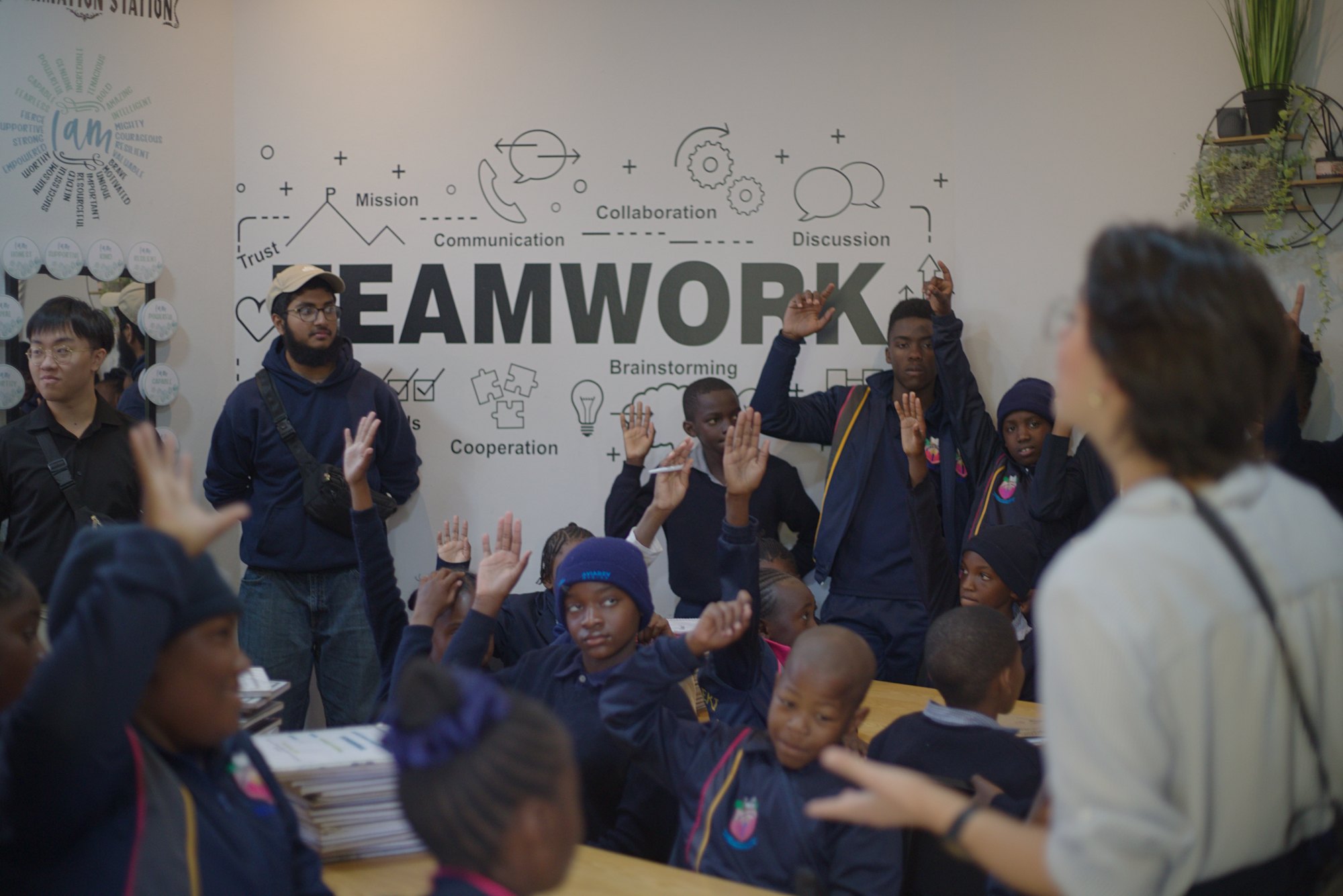 A Namibian classroom full of students raising their hands, with ETHS students standing along the back wall under a TEAMWORK mural